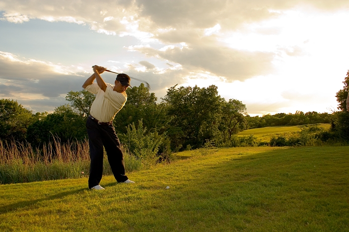 An outdoor sports photo of a golfer at the end of his swing. He is looking toward the setting sun and has scenic green grass, trees and satiny clouds around him.